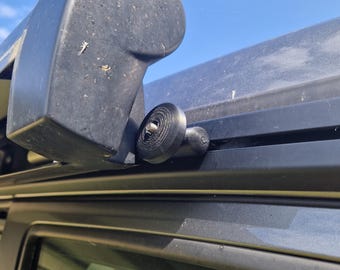 Op de afbeelding: Een zwarte luifelarm met een zwarte kunststof knop en een metalen rail. De luifelarm is bevestigd aan een voertuig.