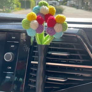 Op de afbeelding: Een handgemaakte auto-ventilatie-decoratie. Het toont een boeket bloemen gemaakt van gekleurd garen, waaronder geel, rood, blauw, groen en paars. De bloemen staan in een zwarte vaas met geometrisch patroon.