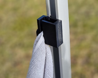 Op de afbeelding: Een zwarte plastic handdoekclip met de tekst "Campfire Kitchen" bevestigd aan een zilveren paal. Een witte handdoek hangt aan de clip.