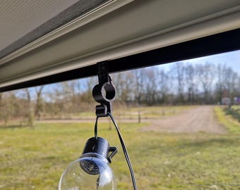 Op de afbeelding: Een zwarte plastic haak met een heldere gloeilamp die eraan hangt. De haak is bevestigd aan een wit zonnescherm. De achtergrond is een wazig beeld van bomen en gras.