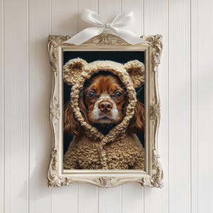 Puede incluir: Retrato enmarcado de un Cavalier King Charles Spaniel con una sudadera con capucha de oso de peluche. El perro tiene pelaje marrón y está centrado en un marco decorativo, adornado y blanquecino. Un lazo blanco está atado en la parte superior del marco.