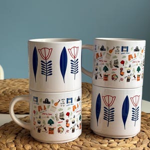 May include: Four white ceramic mugs stacked in two pairs. Two mugs feature a repeating pattern of blue leaves and red-outlined flowers. The other two mugs display a colourful collage of small illustrations, including musical instruments, food, and text.