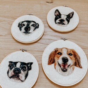 May include: Four round embroidery hoops featuring detailed portraits of dogs. The dogs have various fur colors, including black, white, brown, and tan. The hoops are set against a light-colored fabric background.