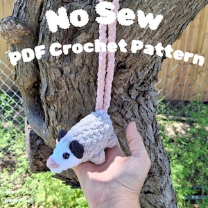 May include: A crocheted opossum toy in gray, white, black, and pink, with the text "No Sew PDF Crochet Pattern" in white. The opossum has a pink nose and a pink cord attached to it.