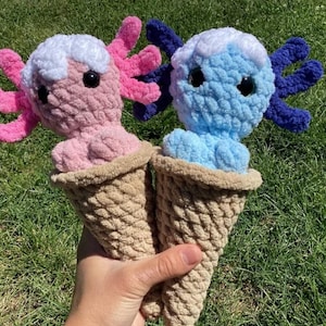 May include: Two handmade crochet axolotl plush toys, one pink and one blue, each nestled in a tan crochet ice cream cone. The axolotls have white and colored frills and black safety eyes. The plushies are held against a backdrop of green grass.