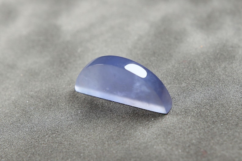 May include: A polished, semi-circular gemstone in a soft blue hue. The cabochon has a smooth, glossy surface with a white, oval-shaped inclusion. The gemstone is set against a gray background.