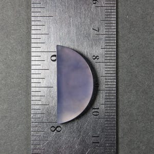 May include: A semi-circular, pale lavender gemstone cabochon, approximately 1.25 inches long, is positioned next to a metal ruler. The ruler shows measurements in inches and fractions of an inch. The gemstone has a smooth, polished surface.