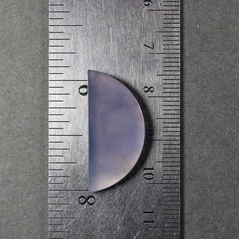 May include: A semi-circular, pale lavender gemstone cabochon, approximately 1.25 inches long, is positioned next to a metal ruler. The ruler shows measurements in inches and fractions of an inch. The gemstone has a smooth, polished surface.