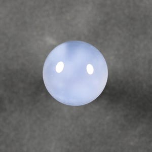 May include: A round, pale blue gemstone with a polished surface. The stone has a translucent quality, with subtle variations in color. Two bright white spots are visible on the surface, reflecting light. The gemstone is set against a gray background.