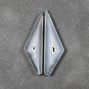 May include: Two matching, light blue and white agate gemstone slices. Each slice is diamond-shaped with a central cavity and a polished surface. The gemstones are displayed against a gray background.