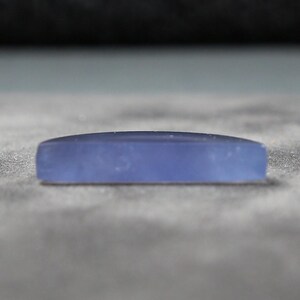 May include: A rectangular, light blue gemstone with a slightly curved top. The stone is smooth and opaque, resting on a textured, gray surface. The gemstone's shape is elongated, with rounded edges.