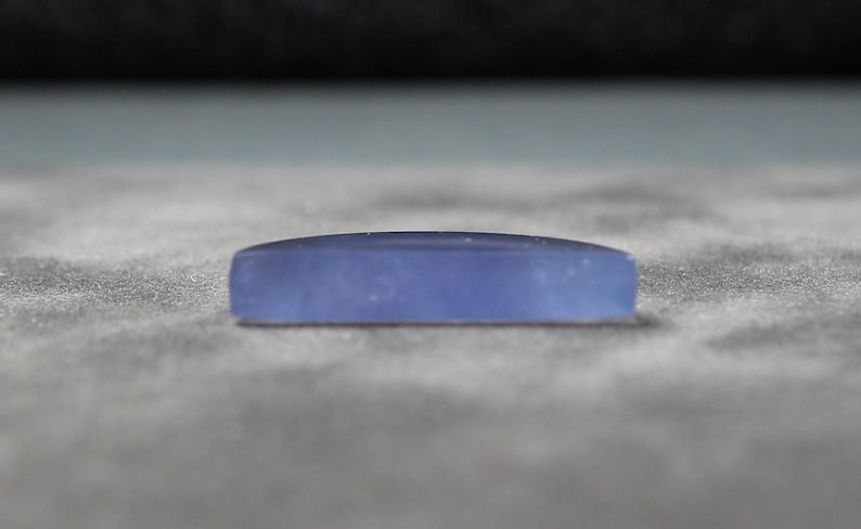 May include: A rectangular, light blue gemstone with a slightly curved top. The stone is smooth and opaque, resting on a textured, gray surface. The gemstone's shape is elongated, with rounded edges.