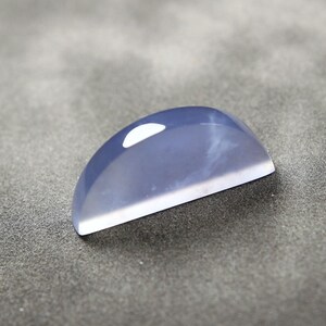 May include: A polished, semi-circular gemstone with a pale blue hue and a white, flat base. The stone has a smooth, reflective surface with a subtle, internal pattern. The gemstone is likely for jewelry making.