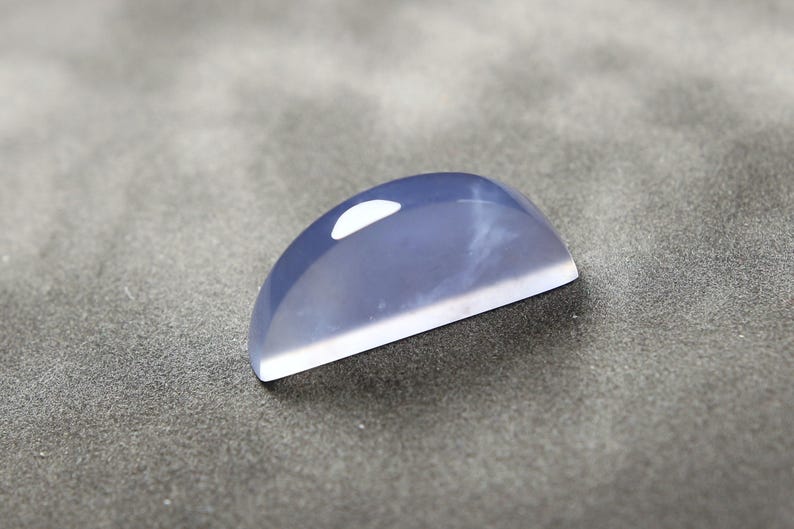 May include: A polished, semi-circular gemstone with a pale blue hue and a white, flat base. The stone has a smooth, reflective surface with a subtle, internal pattern. The gemstone is likely for jewelry making.