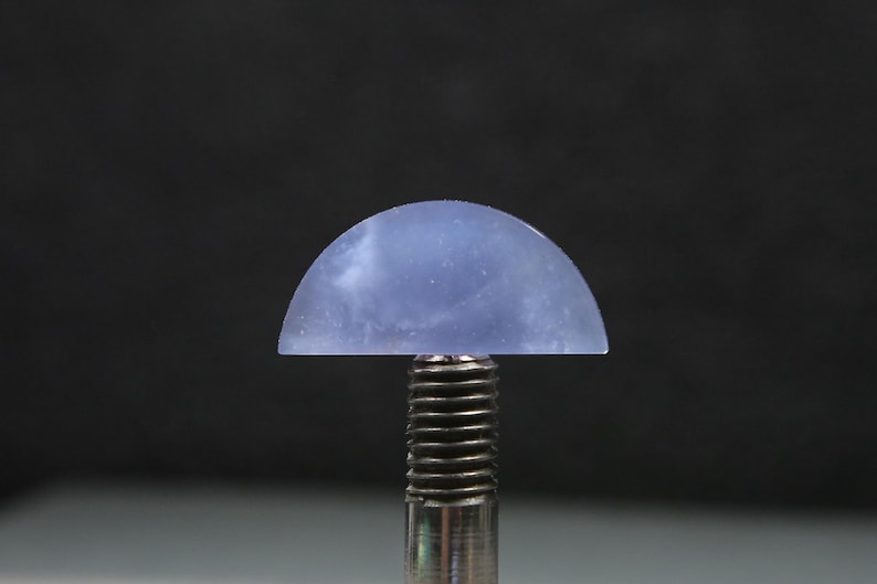 May include: A translucent, half-dome shaped gemstone in shades of blue and white, mounted on a silver metal post. The gemstone has a smooth, polished surface and is set against a dark background. The overall effect is one of elegance and simplicity.
