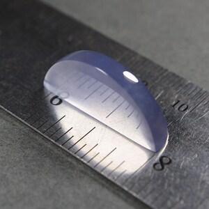 May include: A translucent, oval-shaped, pale blue gemstone is positioned on a metal ruler. The gemstone is approximately 1 inch in length. The ruler's markings are clearly visible, providing a size reference.