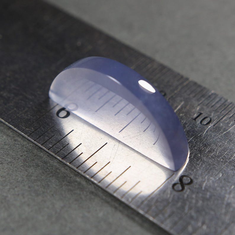 May include: A translucent, oval-shaped, pale blue gemstone is positioned on a metal ruler. The gemstone is approximately 1 inch in length. The ruler's markings are clearly visible, providing a size reference.