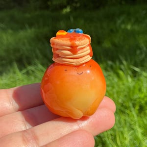 May include: A miniature orange fruit with a smiling face, topped with a stack of pancakes drizzled with syrup and decorated with blueberries and orange slices. The miniature food item is held in a hand.