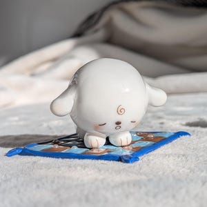 May include: A small white ceramic figurine of a puppy with closed eyes and a brown swirl on its forehead, resting on a square blue, brown, and light blue checkered mat with a raised blue border and small raised dots on the surface.