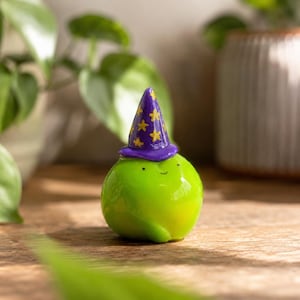 Può includere: Figurina verde lucida di una creatura rotonda con un cappello da mago viola ornato di stelle dorate. La figurina ha un piccolo viso sorridente. Lo sfondo comprende piante verdi e una superficie in legno.