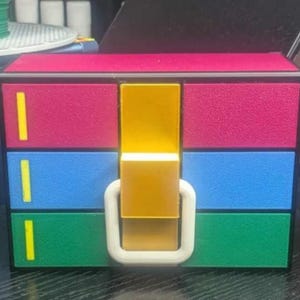 May include: A small, colourful storage box with a hinged lid and a white handle. The box has three drawers, each with a different colour: red, blue, and green. Each drawer has a yellow stripe on the front.