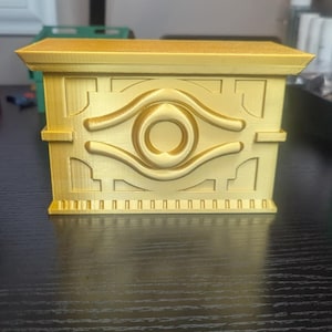 May include: A gold-colored, 3D printed box with a raised design of the Eye of Horus. The box has a detailed, intricate design and a textured surface.