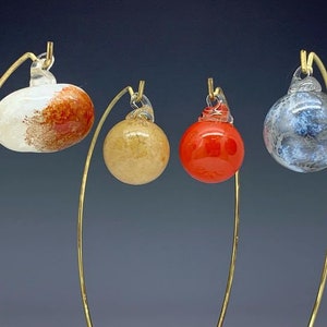 Puede incluir: Cuatro adornos de vidrio colgados de soportes de metal dorado. Los adornos son un remolino blanco y naranja, un remolino tostado, un remolino rojo y un remolino azul y gris.