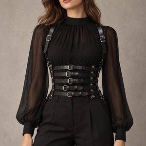 Op de afbeelding: Een zwarte blouse met lange transparante mouwen en een hoge halslijn, gedragen onder een zwart lederlook korset-harnas met vijf horizontale banden met zilverkleurige metalen gespen en klinknagels, gecombineerd met een zwarte pantalon.