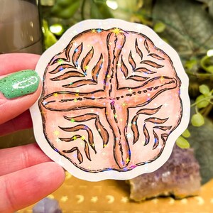 May include: A round sticker with a light brown design featuring a cross and leaf-like patterns. The sticker has a holographic, iridescent border. The background includes greenery, a candle, and crystals.