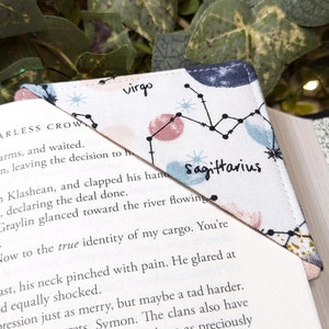 May include: A white fabric bookmark with a pink and blue constellation design. The bookmark features the zodiac signs Virgo and Sagittarius.