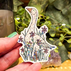 May include: A sticker featuring a white goose with a long neck, standing among purple and pink flowers and green grass. The goose has a smiling expression. The sticker has a holographic effect.