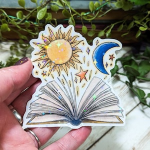 May include: A holographic sticker featuring an open book with a sun and moon above it. The sun and moon are surrounded by stars. The sticker is on a wooden surface.