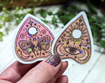 Può includere: Due adesivi per planchette di tavoletta Ouija olografici rosa e arancioni. Uno presenta una falena con una luna crescente e stelle. L'altro presenta un sole, un occhio e una luna crescente.