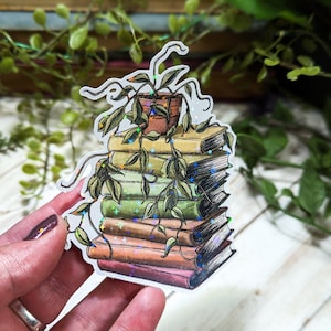 May include: A sticker of a stack of books with a potted plant growing on top. The books are various colors and have a glittery finish. The plant is green with a brown pot.