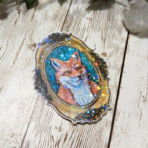 K&ouml;nnte beinhalten: Ein holografischer Aufkleber mit einem roten Fuchs in einem goldenen, verzierten Rahmen vor einem blauen Sternenhimmel.