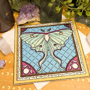 Peut inclure: Impression d'art carr&eacute;e repr&eacute;sentant un papillon lune. Le papillon a des ailes vert p&acirc;le avec des accents roses et violets, sur un fond bleu clair &agrave; motif de losanges. L'impression est encadr&eacute;e d'une bordure jaune et d'&eacute;l&eacute;ments d&eacute;coratifs.