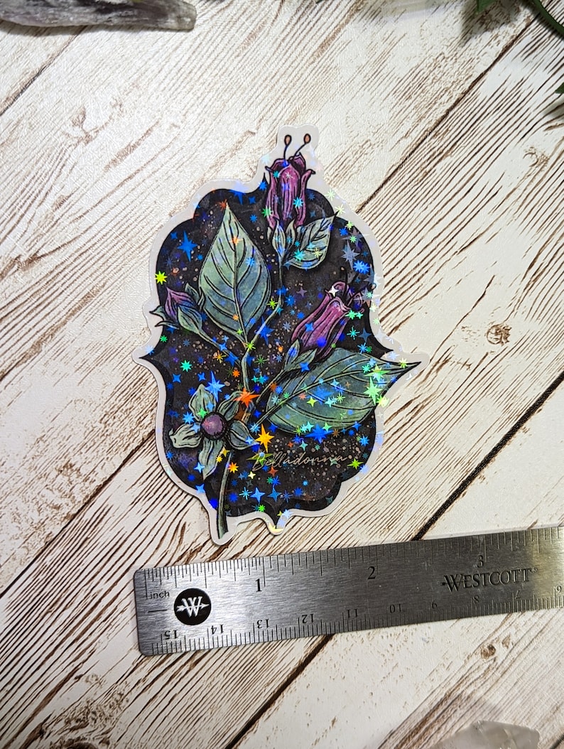 May include: A holographic sticker featuring a purple flower with a dark background and a scattering of iridescent stars. The sticker is approximately 5 cm in diameter.