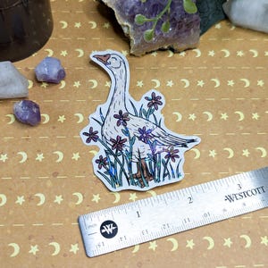 May include: A sticker featuring a white goose with a long neck, surrounded by purple and pink flowers. The goose has a light orange beak and feet. A silver ruler with inch measurements is next to the sticker. The background is tan with a crescent moon and star pattern.