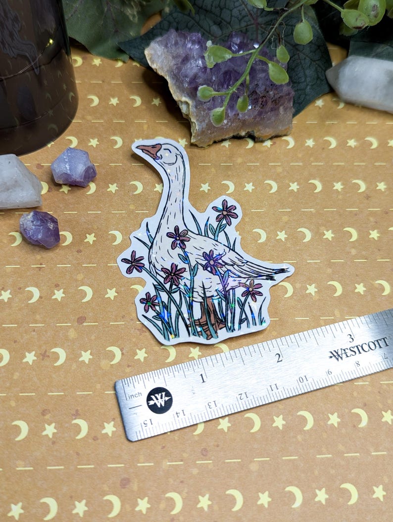 May include: A sticker featuring a white goose with a long neck, surrounded by purple and pink flowers. The goose has a light orange beak and feet. A silver ruler with inch measurements is next to the sticker. The background is tan with a crescent moon and star pattern.