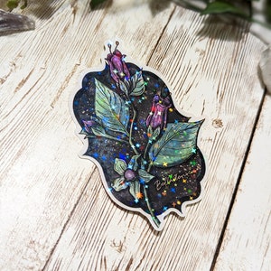 May include: A holographic sticker featuring a purple flower with green leaves and a black background. The sticker has a glittery finish and the text "Bellatonna" is written on it.
