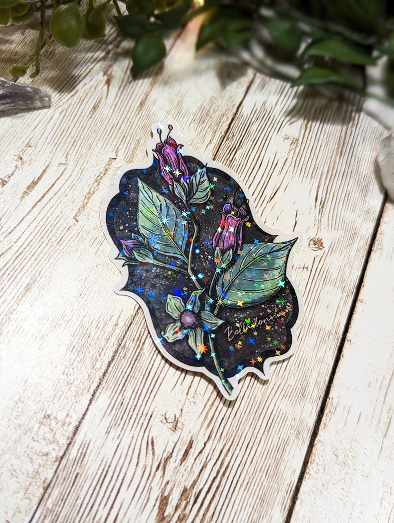 May include: A holographic sticker featuring a purple flower with green leaves and a black background. The sticker has a glittery finish and the text "Bellatonna" is written on it.