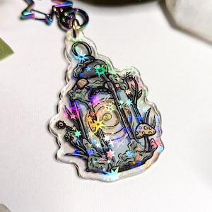 May include: A holographic charm featuring a lantern with a spiral design inside, surrounded by stars, mushrooms, and foliage. The charm is attached to a silver star-shaped keychain.