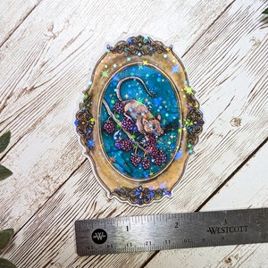 May include: A holographic sticker featuring a detailed illustration of a rat climbing a branch with berries. The rat is brown and white, and the berries are purple. The background is a teal blue with gold stars and a gold ornate frame.