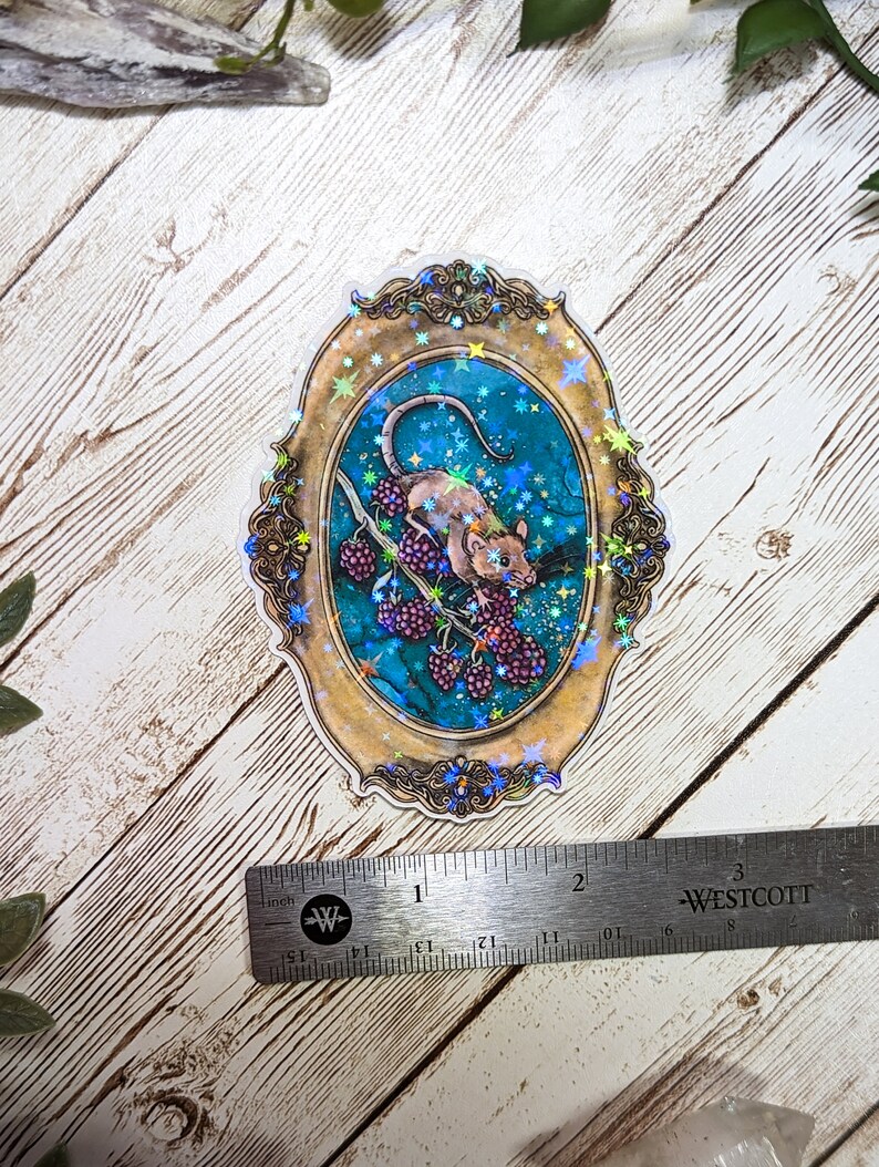 May include: A holographic sticker featuring a detailed illustration of a rat climbing a branch with berries. The rat is brown and white, and the berries are purple. The background is a teal blue with gold stars and a gold ornate frame.