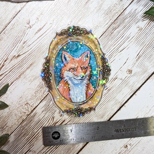 K&ouml;nnte beinhalten: Ein holografischer Aufkleber mit einem Fuchs in einem goldenen verzierten Rahmen auf blauem Hintergrund mit goldenen Sternen.