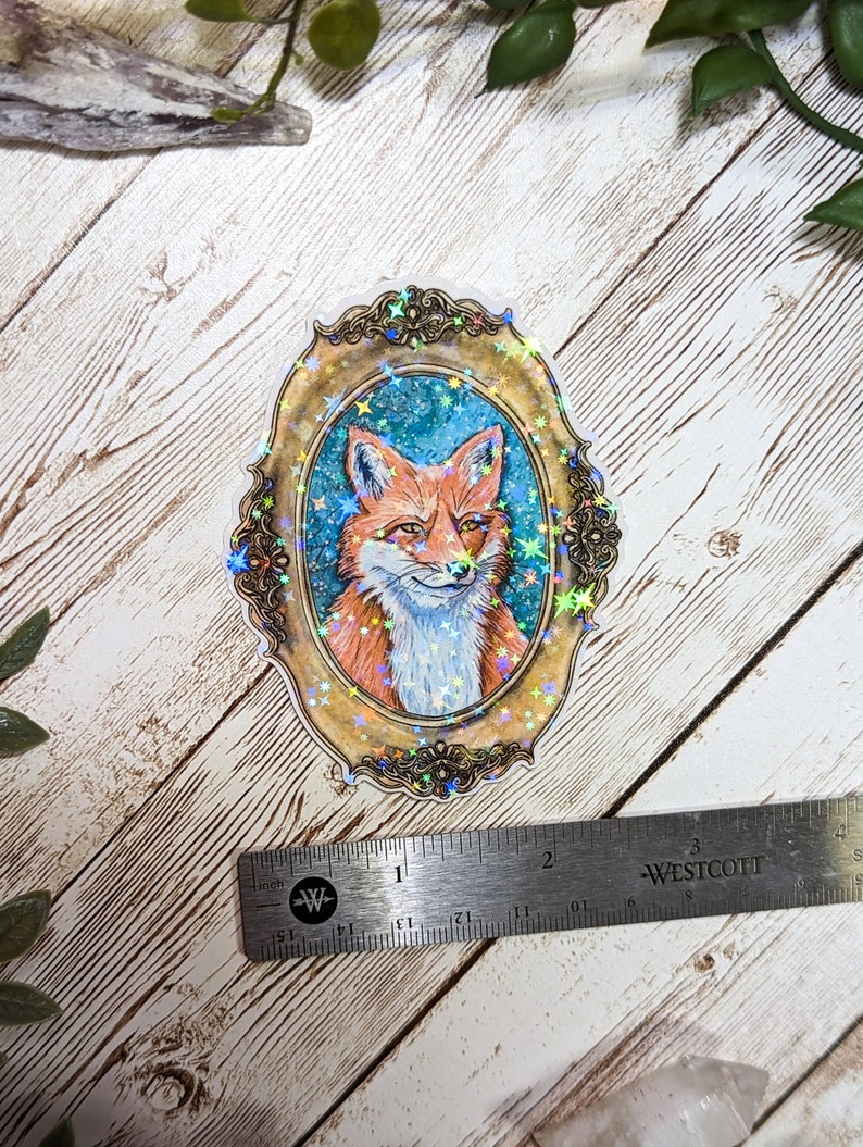 K&ouml;nnte beinhalten: Ein holografischer Aufkleber mit einem Fuchs in einem goldenen verzierten Rahmen auf blauem Hintergrund mit goldenen Sternen.