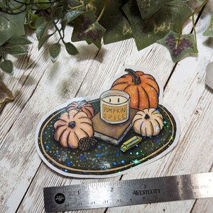 May include: A sticker featuring a hand-drawn illustration of a candle, pumpkins, and a book on a plate with a glittery background. The candle is labeled "Pumpkin Spice".