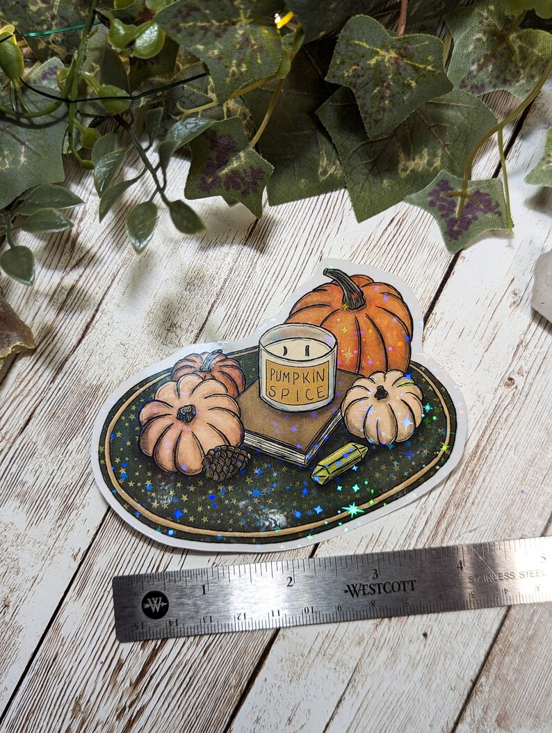 May include: A sticker featuring a hand-drawn illustration of a candle, pumpkins, and a book on a plate with a glittery background. The candle is labeled "Pumpkin Spice".