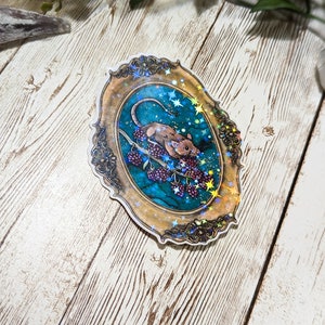 May include: A sticker featuring a detailed illustration of a mouse eating berries on a vine, set against a starry night sky. The image is framed in a gold, ornate oval frame with a holographic finish.