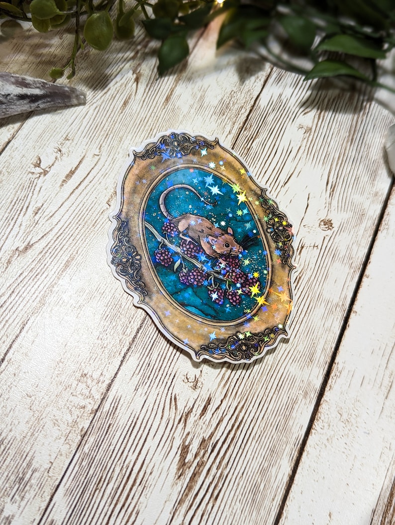 May include: A sticker featuring a detailed illustration of a mouse eating berries on a vine, set against a starry night sky. The image is framed in a gold, ornate oval frame with a holographic finish.